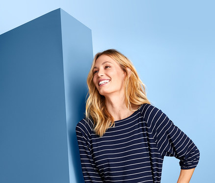 Mosolygó nő kék falnak dőlve, denevérujjú felsőt és fehér nadrágot visel.