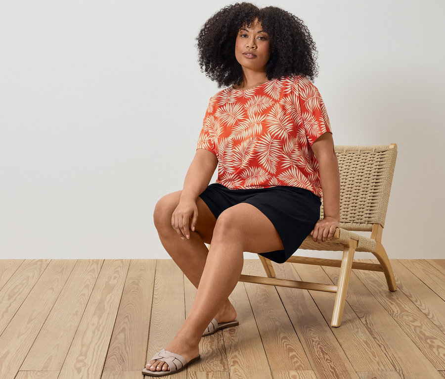 Egy nő ül egy széken, nyomott mintás női pólóblúzt és fekete jersey rövidnadrágot visel.