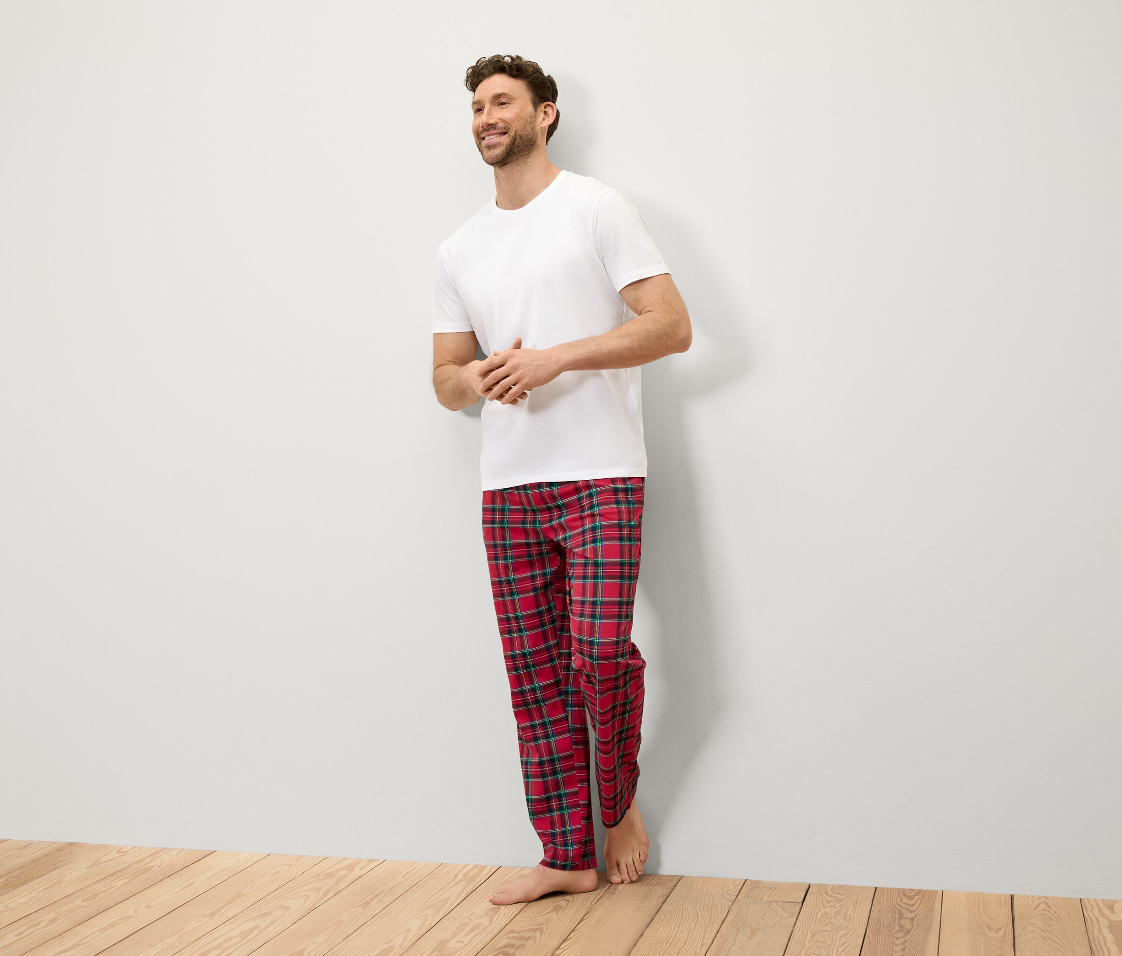 Egy mosolygó férfi áll a falnál, fehér pólót és piros kockás nadrágot visel.