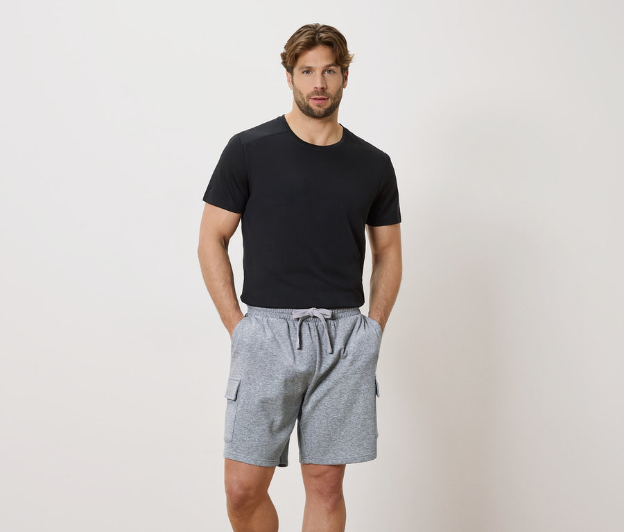 Egy férfi kezével egy szürke, húzózsinóros rövidnadrág zsebében és egy fekete, rövid ujjú pólóban.