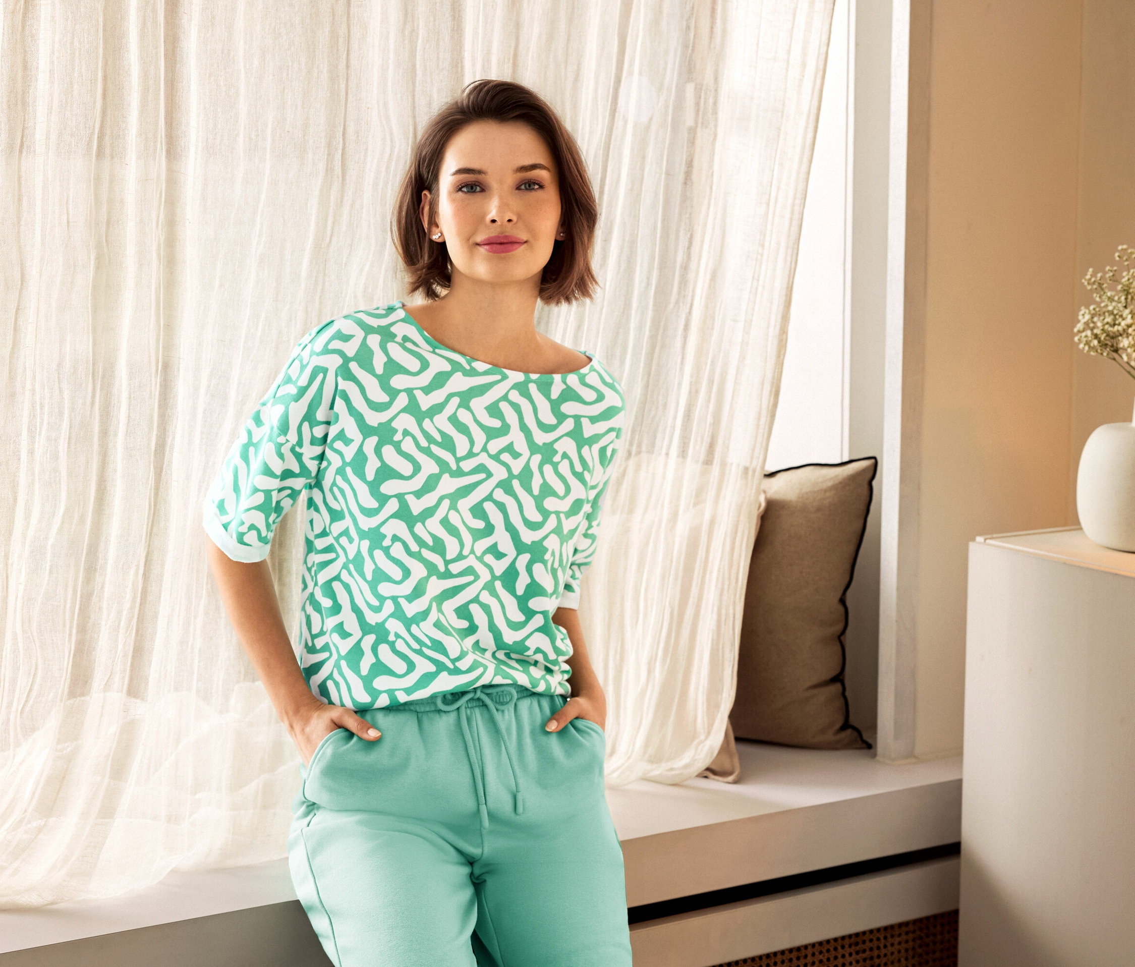 Nő zöld nadrágban és mintás felsőben pózol egy ablak mellett. A háttérben egy kerámia váza látható.