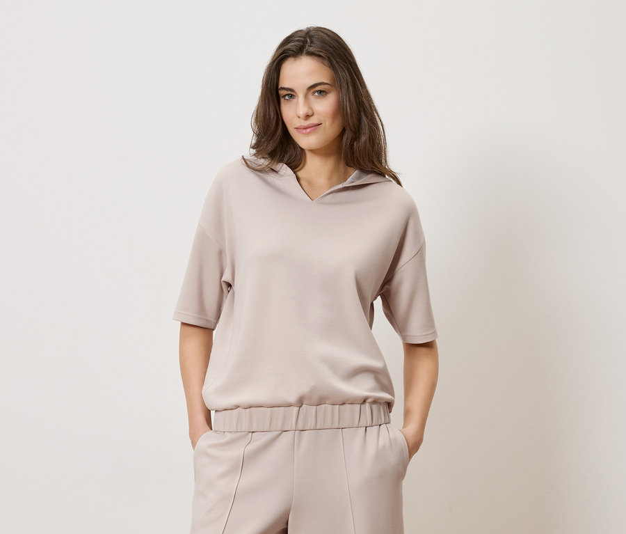 Barna hajú nő bézs kapucnis felsőt és hozzáillő nadrágot visel egy egyszerű háttér előtt.