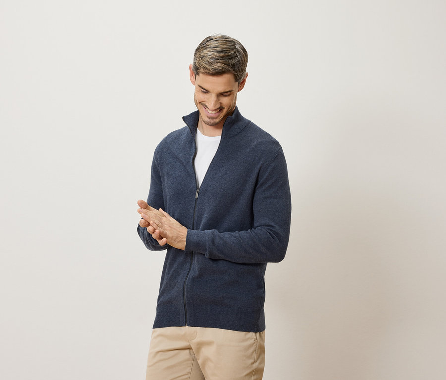 Egy férfi mosolyogva dörzsöli a kezét. Sötétkék kardigánt és bézs nadrágot visel.