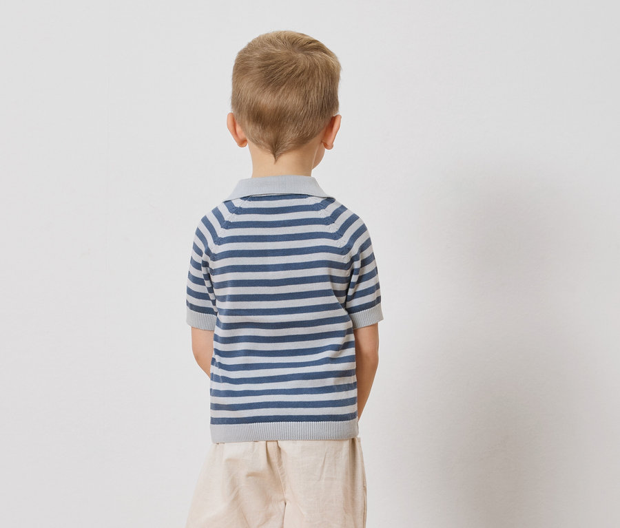 Hátulról látható egy szőke hajú gyermek, aki kék-fehér csíkos pólót és bézs színű rövidnadrágot visel egy fehér háttér előtt.