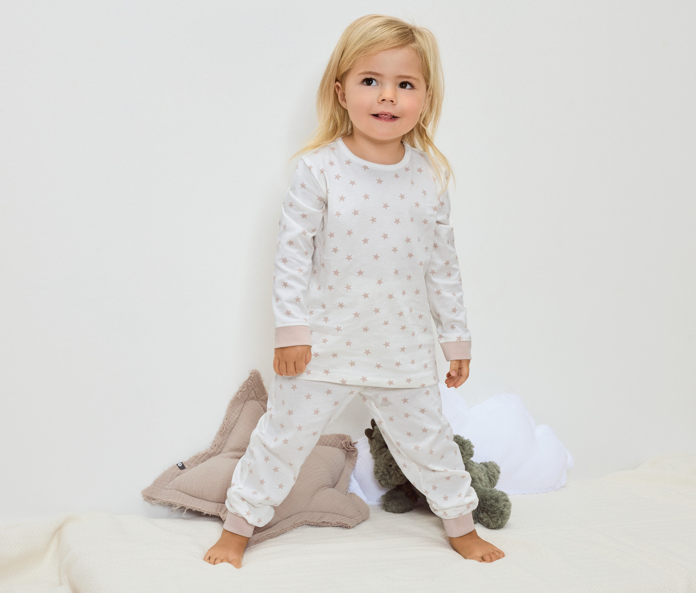 Egy kis szőke lány mezítláb áll egy takarón, csillagmintás fehér pizsamában.