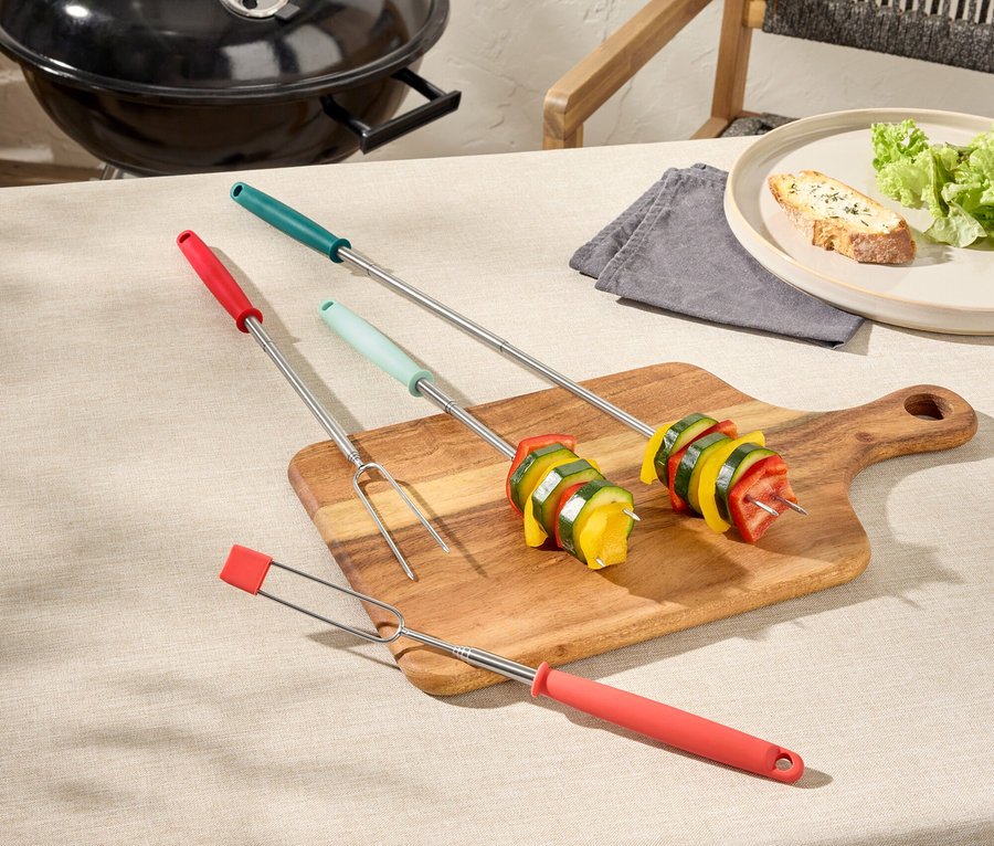 Három zöldséges grillnyárs egy fa deszkán, egy grill és egy tányér kenyér a háttérben.