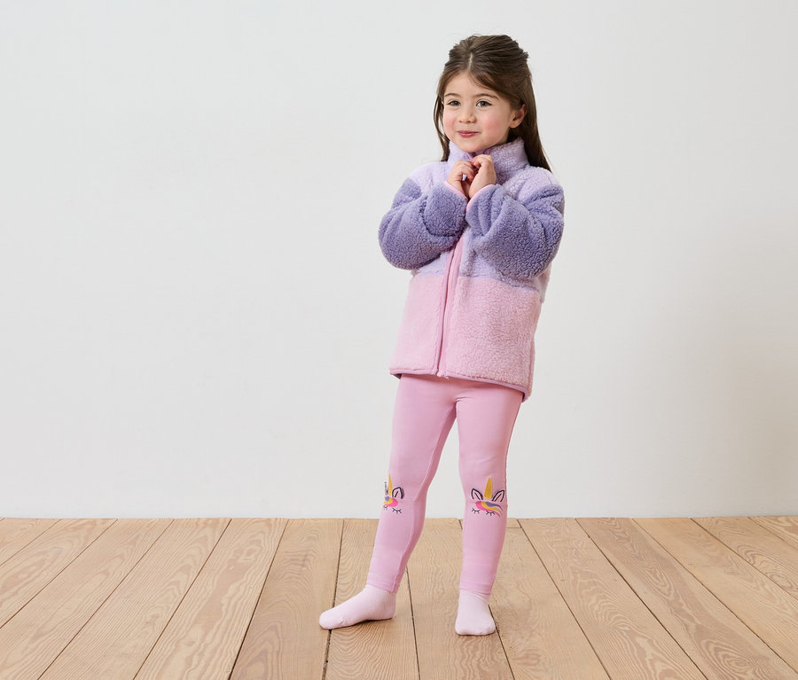 Egy lila-rózsaszín polárkabátot és egyszarvú mintás rózsaszín leggingset viselő lány áll egy fapadlón egy fehér fal előtt.