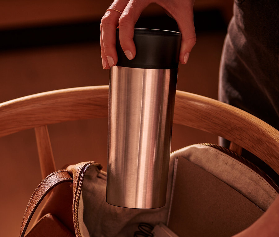 Egy kéz ezüst Coffee-to-go bögrét tesz egy nyitott barna bőrtáskába egy fa széken.