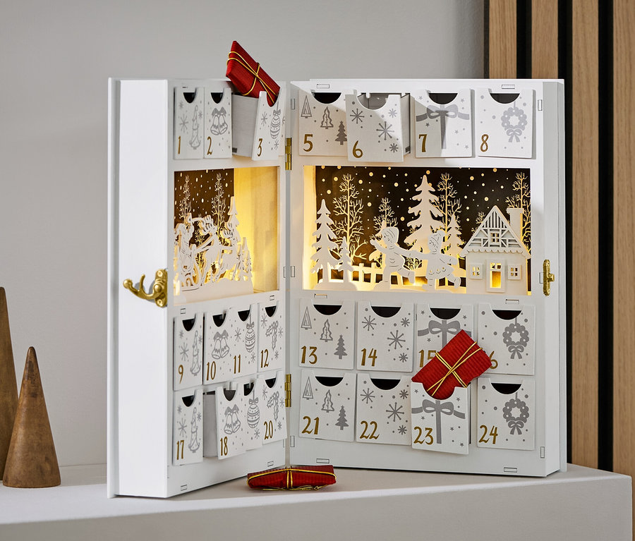 Nyitott, fehér adventi naptár díszekkel és ajándékokkal, három fa kúp mellett.