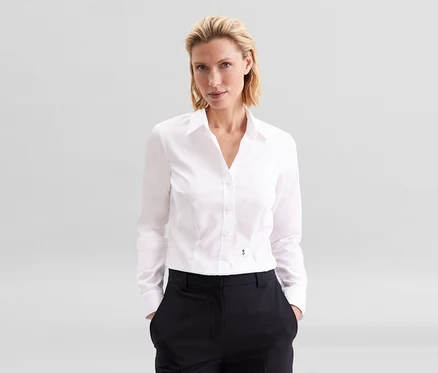 Szőke nő áll a zsebében a kezével, fehér Seidensticker slim fit szabású vasalást nem igénylő ingblúzban és sötét nadrágban.
