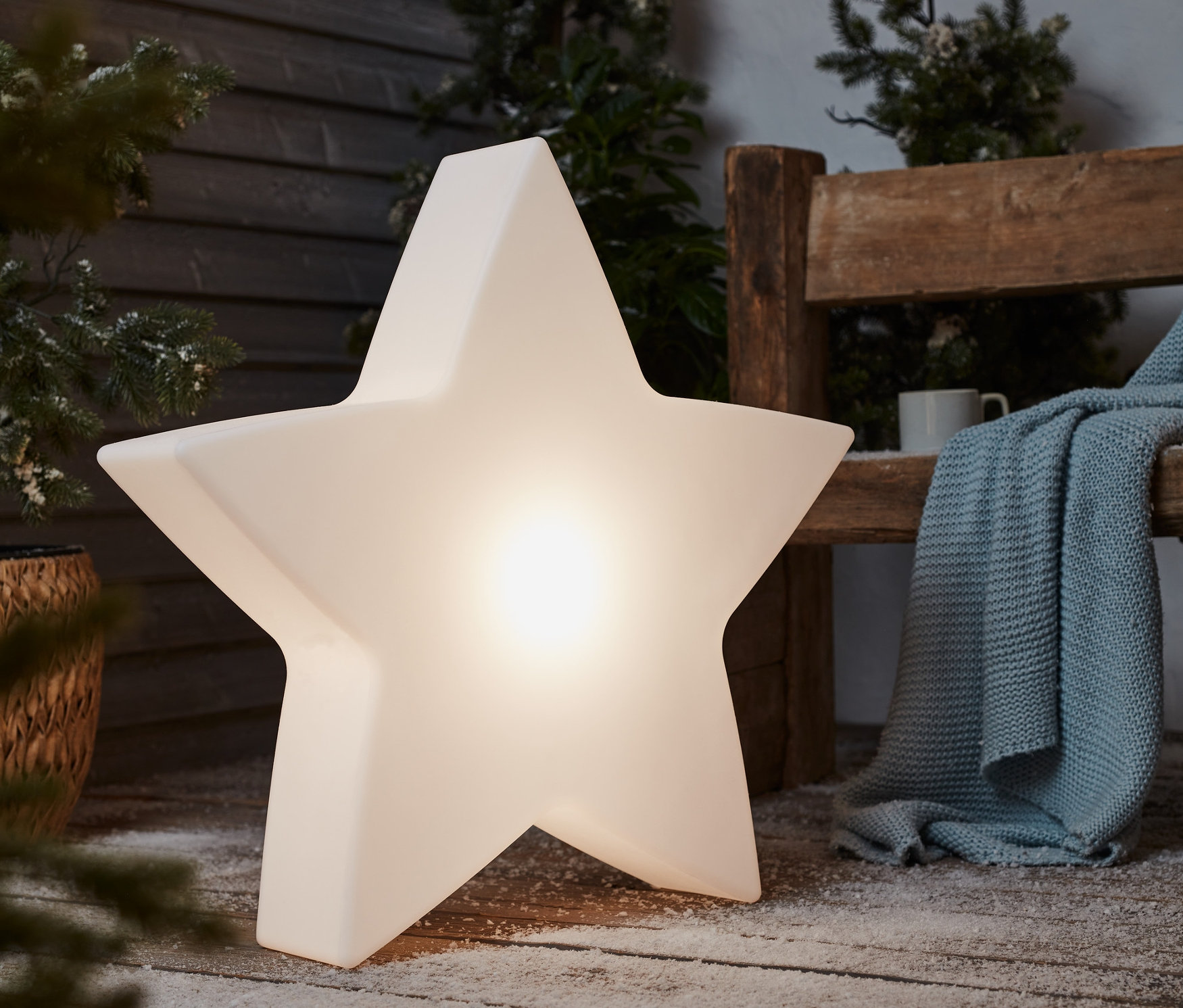 Egy nagy, csillag alakú napelemes kerti lámpa áll egy havas, fa teraszon egy pad mellett.