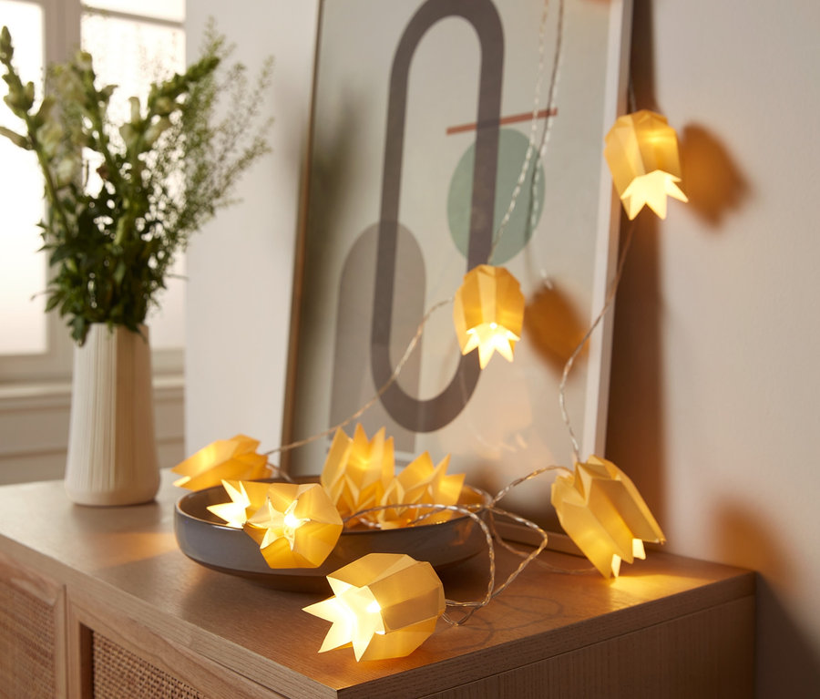 Egy LED-es papír égősor egy fa komódon fekszik egy virágváza mellett.