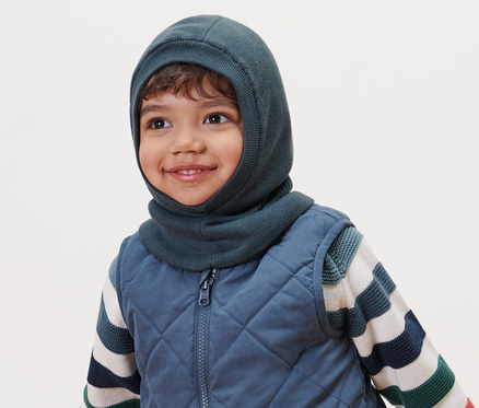 Mosolygó kisfiú kisgyerek csősapkát, steppelt mellényt és csíkos pulóvert visel.