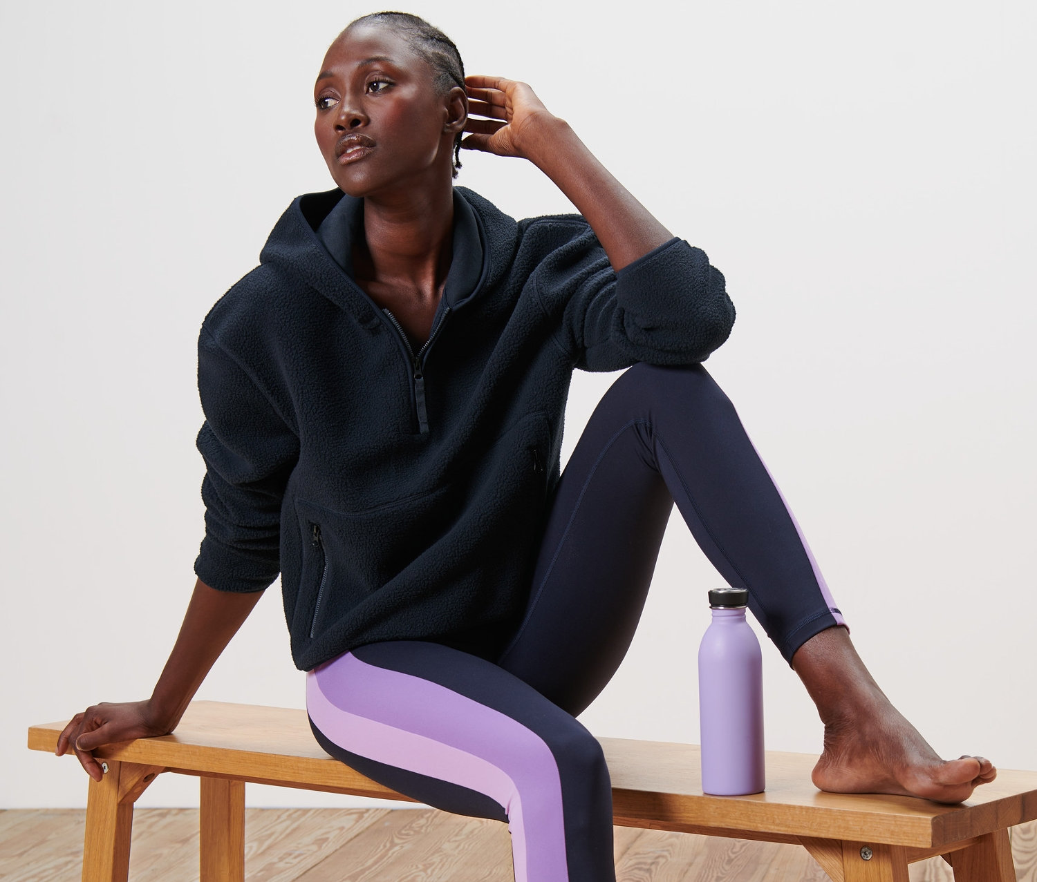 Nő ül egy padon sötétkék plüss kapucnis pulóverben és lila csíkos leggingsben. Mellette egy lila vizespalack.