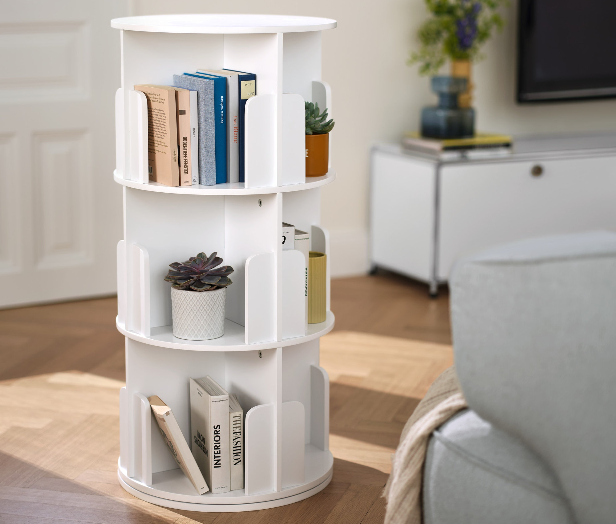 Előtérben forgatható könyvespolc könyvekkel és növényekkel. A háttérben alacsony komód »CN3« fiókokkal, fehér.