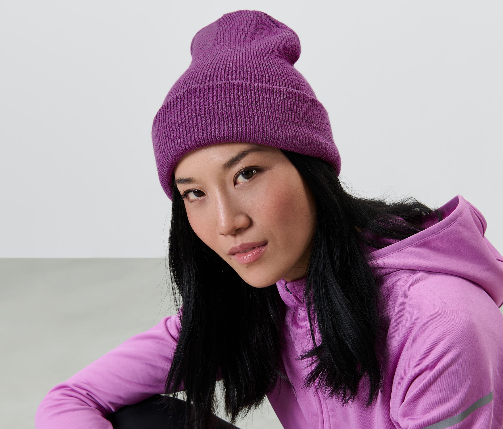 Egy ázsiai nő hosszú, fekete hajjal lila kötött sapkát és lila kapucnis pulóvert visel.
