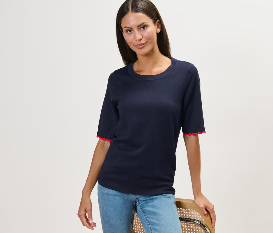 Nő pózol egy széken, sötétkék, finom kötésű pulóvert és farmert visel.