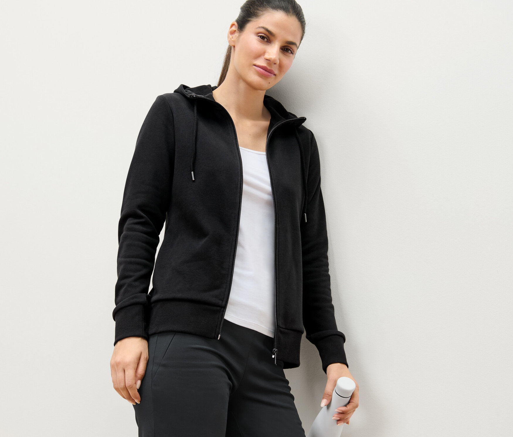 A falnak támaszkodó nő fekete szabadidőfelsőt visel, és egy fehér vizes palackot tart.