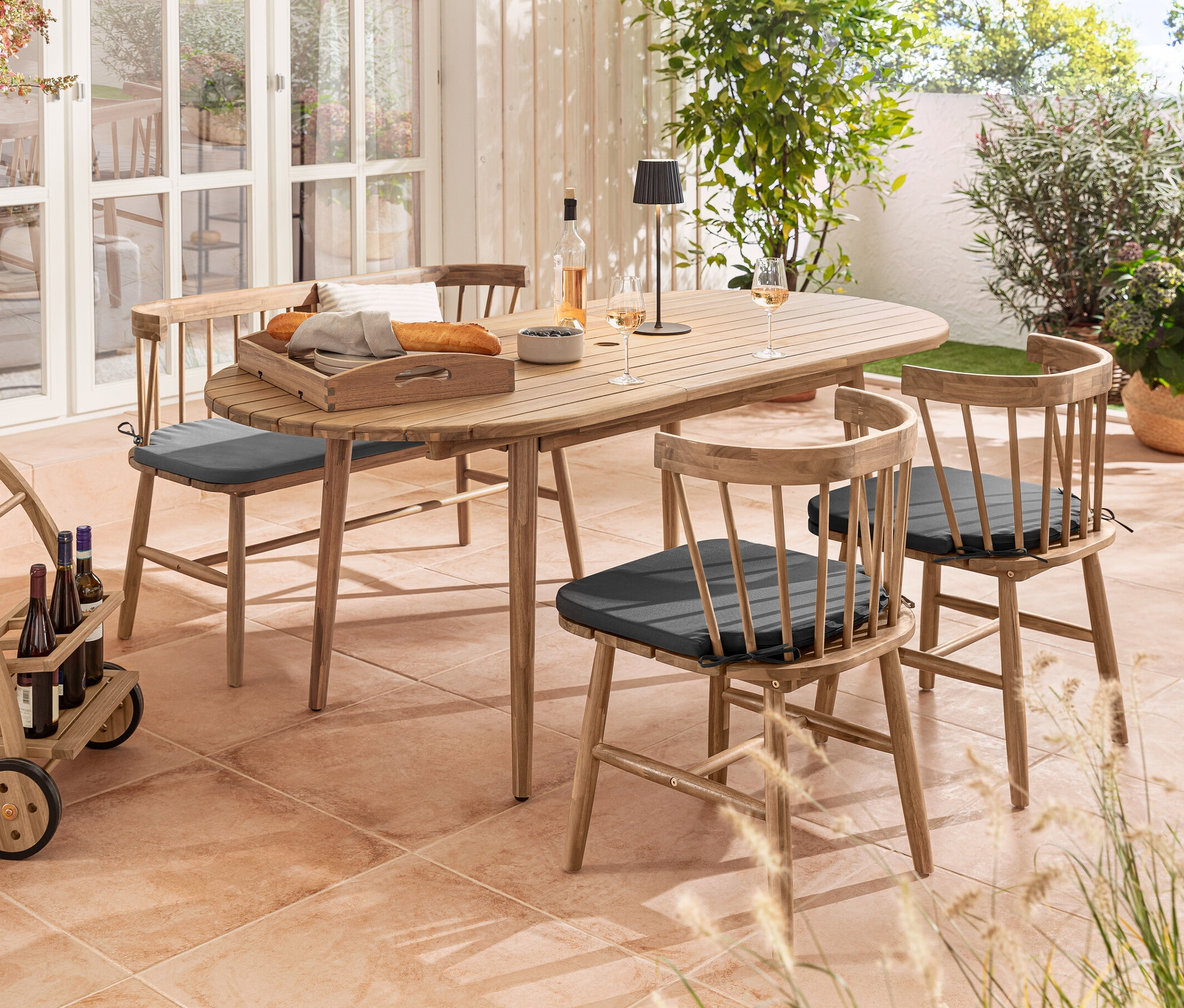 Kültéri jelenet kihúzható ovális asztallal, étkezőszékek üléspárnával, pad ülőpárnával, zsúrkocsi és fekete LED-es akkumulátoros kültéri asztali lámpa.