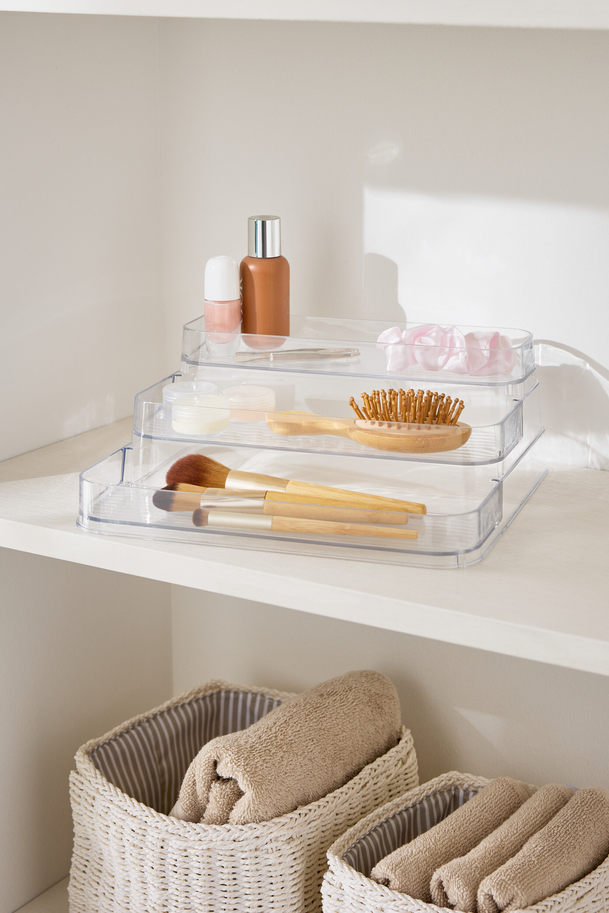 Háromszintes, átlátszó sminkrendszerező egy fehér polcon, törölközőkosarak mellett.