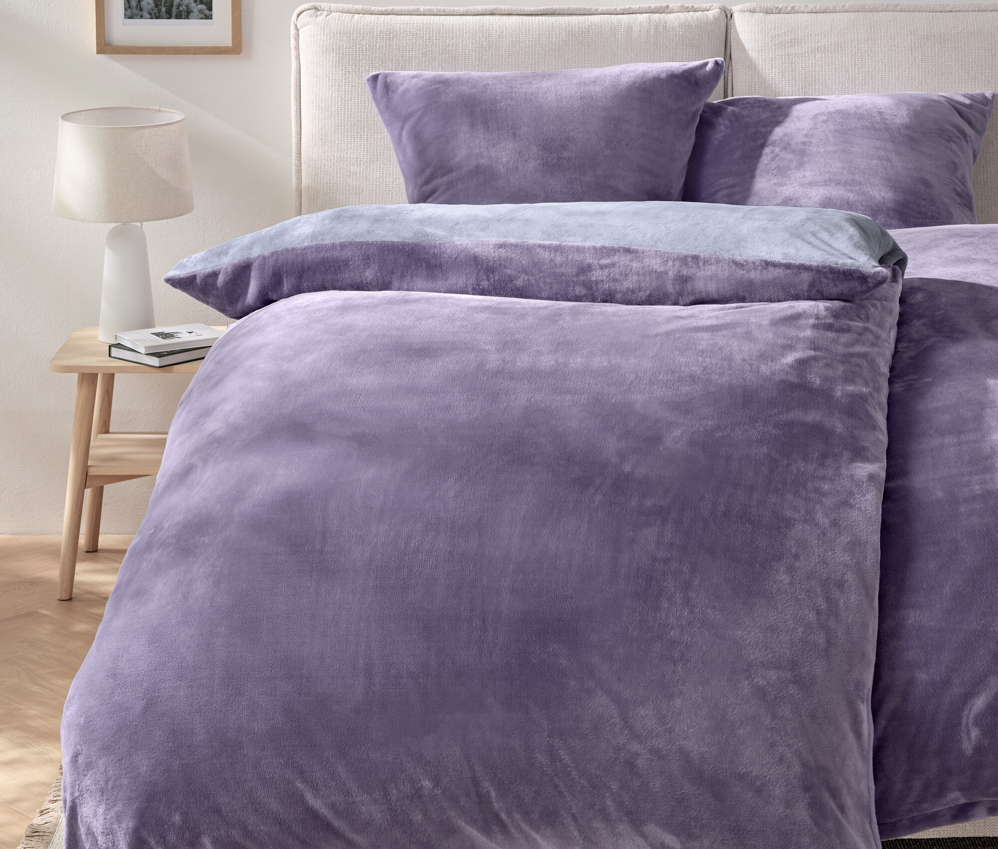 Lila ágynemű egy ágyon két párnával és egy lámpával ellátott éjjeliszekrénnyel.