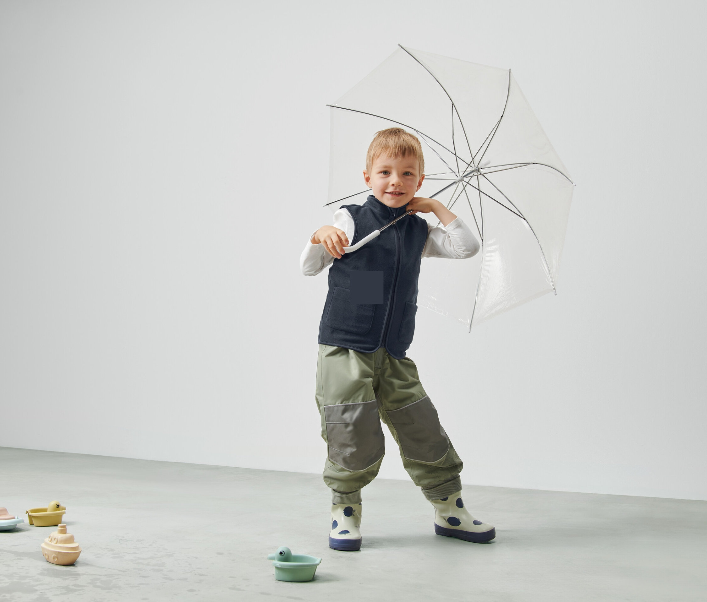 Egy fiú átlátszó esernyőt tart, mellényt és nadrágot visel, és egy szobában áll.