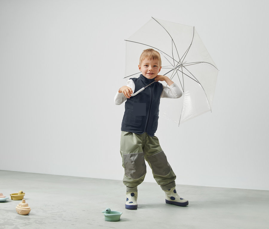 Egy fiú átlátszó esernyőt tart, mellényt és nadrágot visel, és egy szobában áll.