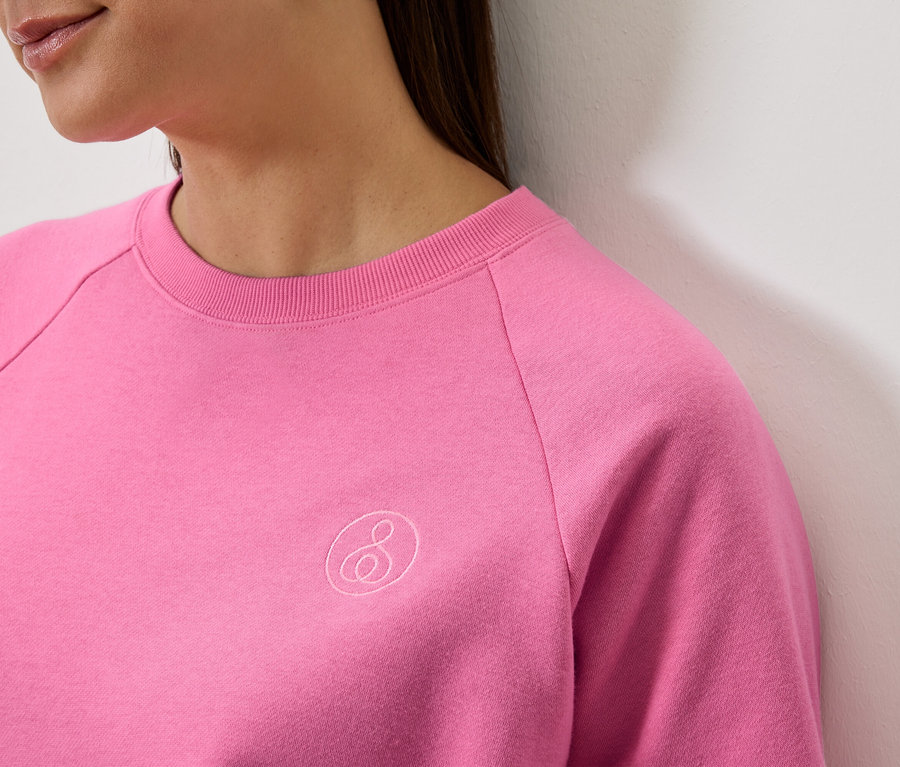 Egy nő részlete, aki rózsaszín, hosszú ujjú pulóvert visel, logóval.