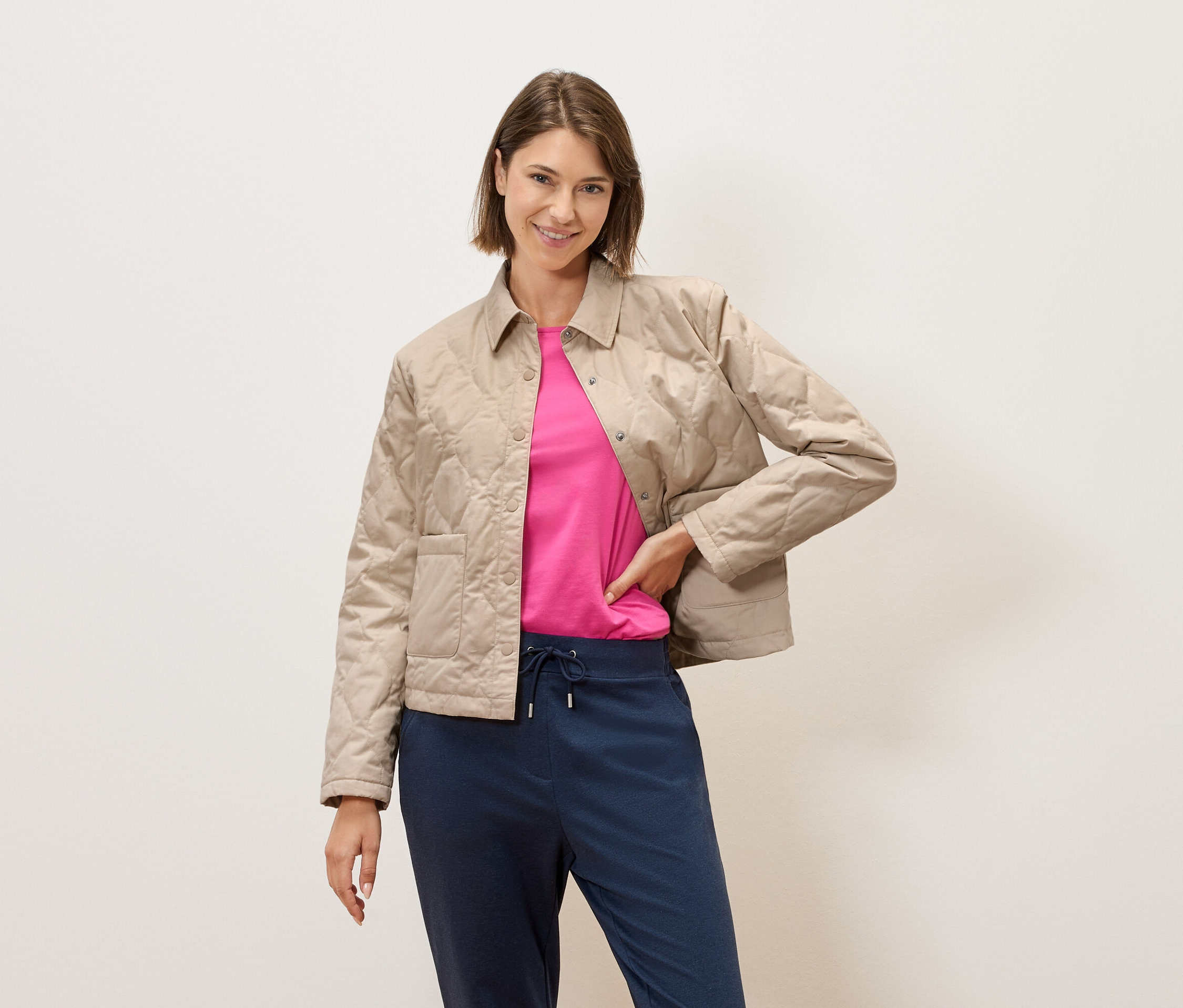 Rövid barna hajú nő pózol egy krémszínű fal előtt, bézs színű steppelt kabátban, rózsaszín pólóban és kék nadrágban.