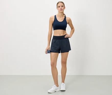 Nő áll csípőre tett kézzel, tengerészkék sportmelltartót, rövidnadrágot és fehér tornacipőt visel, és egy vizespalackot tart.