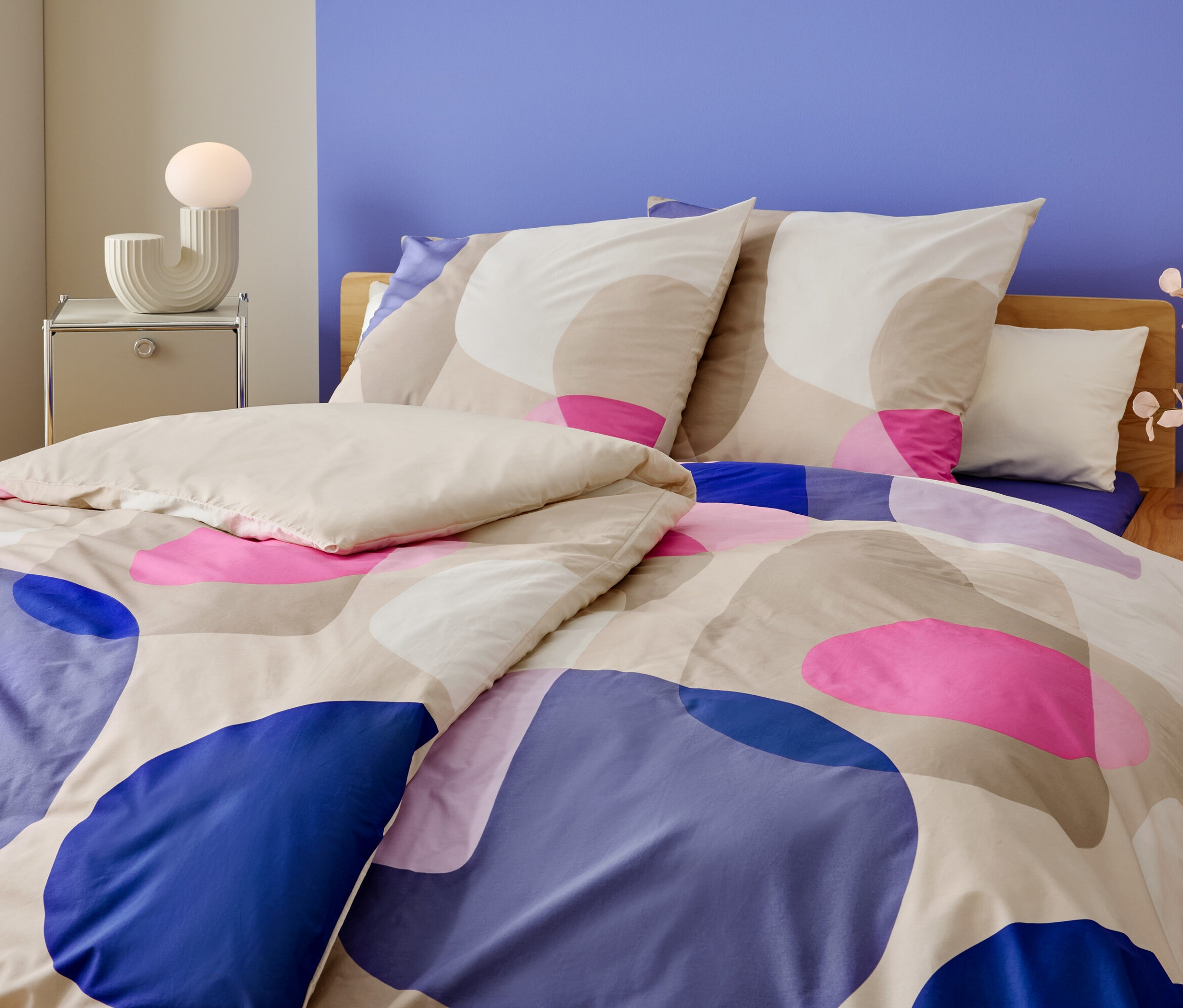 Egy ágy közeli képe kék, rózsaszín és bézs színű geometriai mintával. Az éjjeliszekrényen egy lámpa és egy váza virág van.