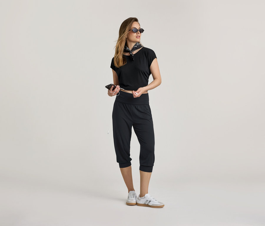 Fekete felsőt és 3/4-es jóganadrágot viselő nő telefonnal és fülhallgatóval. Kendőt és tornacipőt visel.