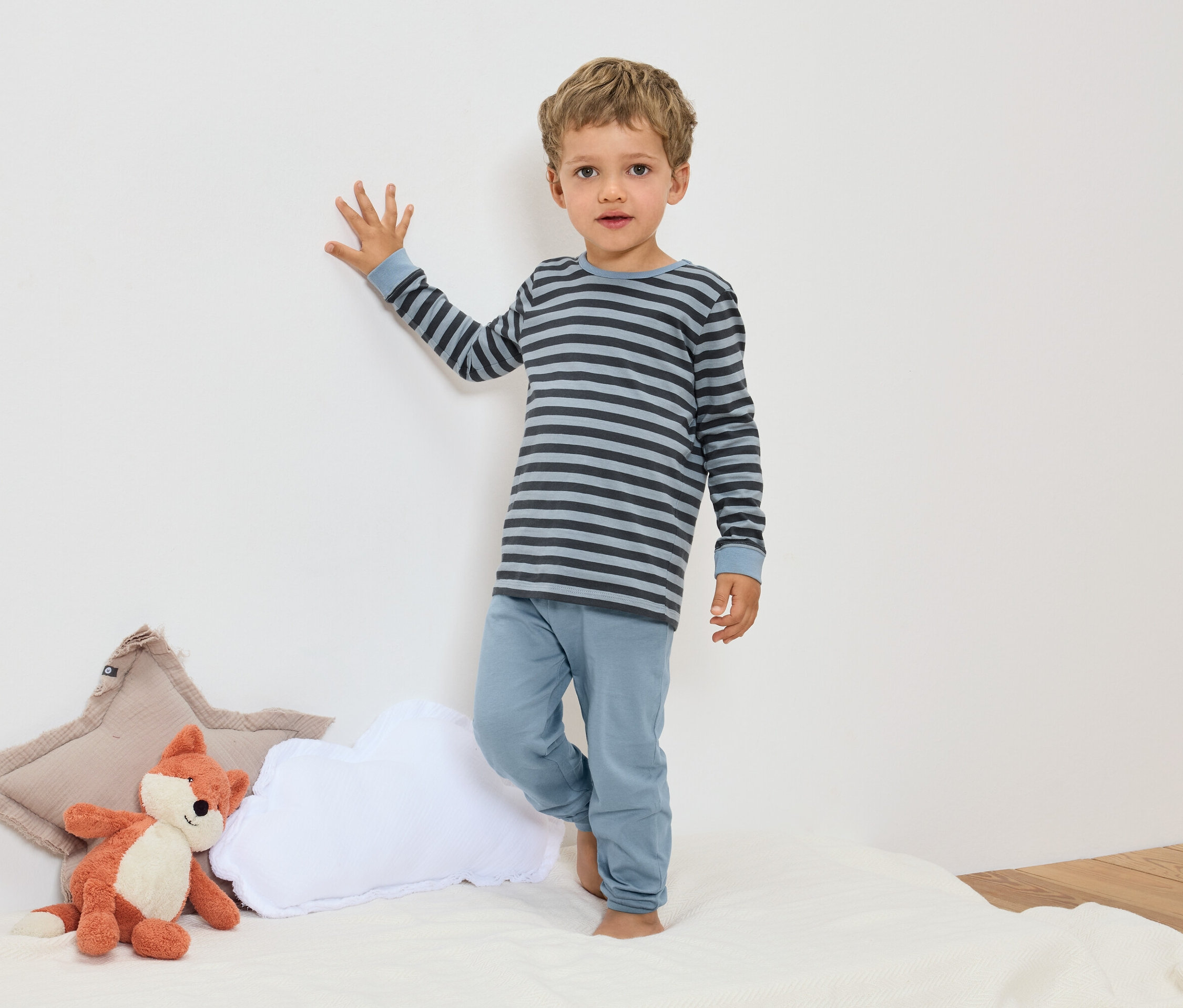 Egy fiú kék csíkos pizsamában áll egy róka plüssállat és párnák mellett.