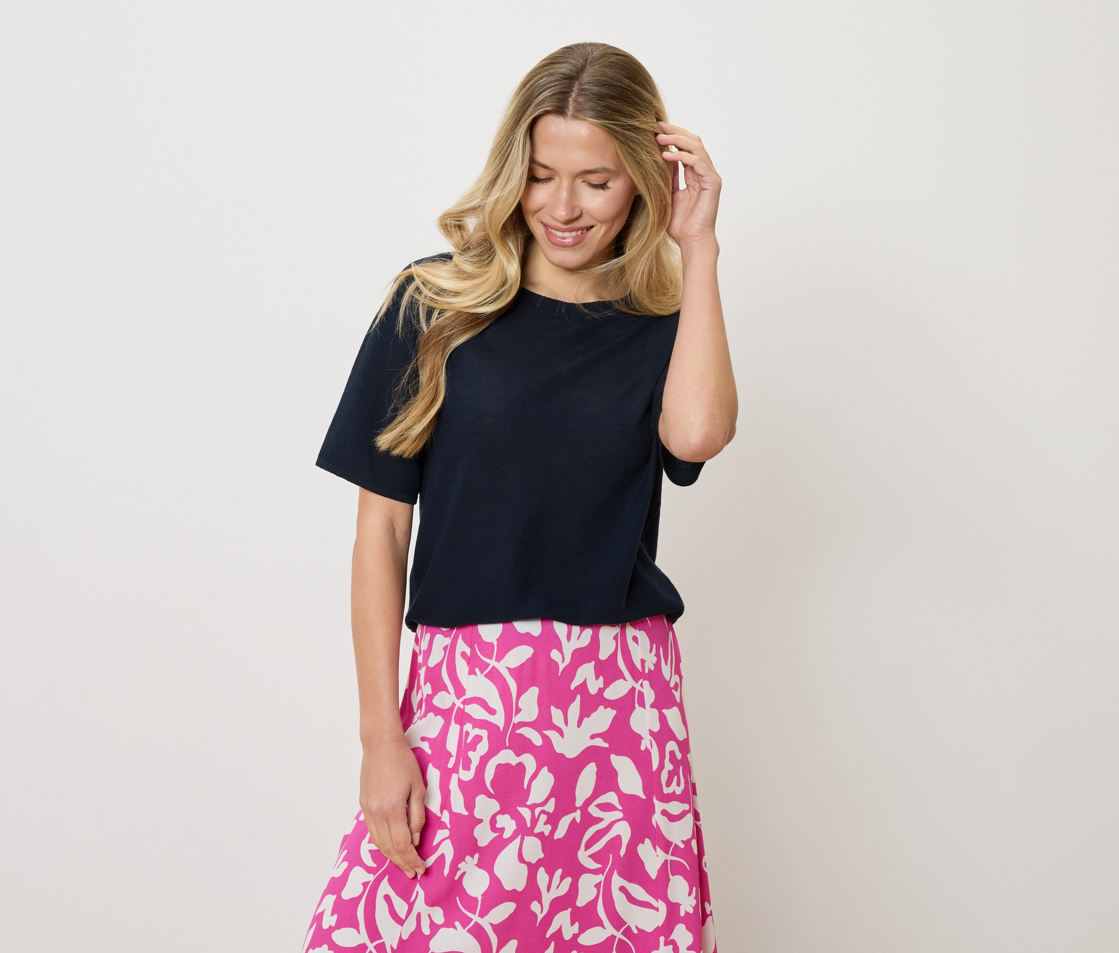 Szőke, hullámos hajú nő, sötétkék pólót és rózsaszín-fehér virágmintás szoknyát visel, és az egyik kezével a haját érinti.