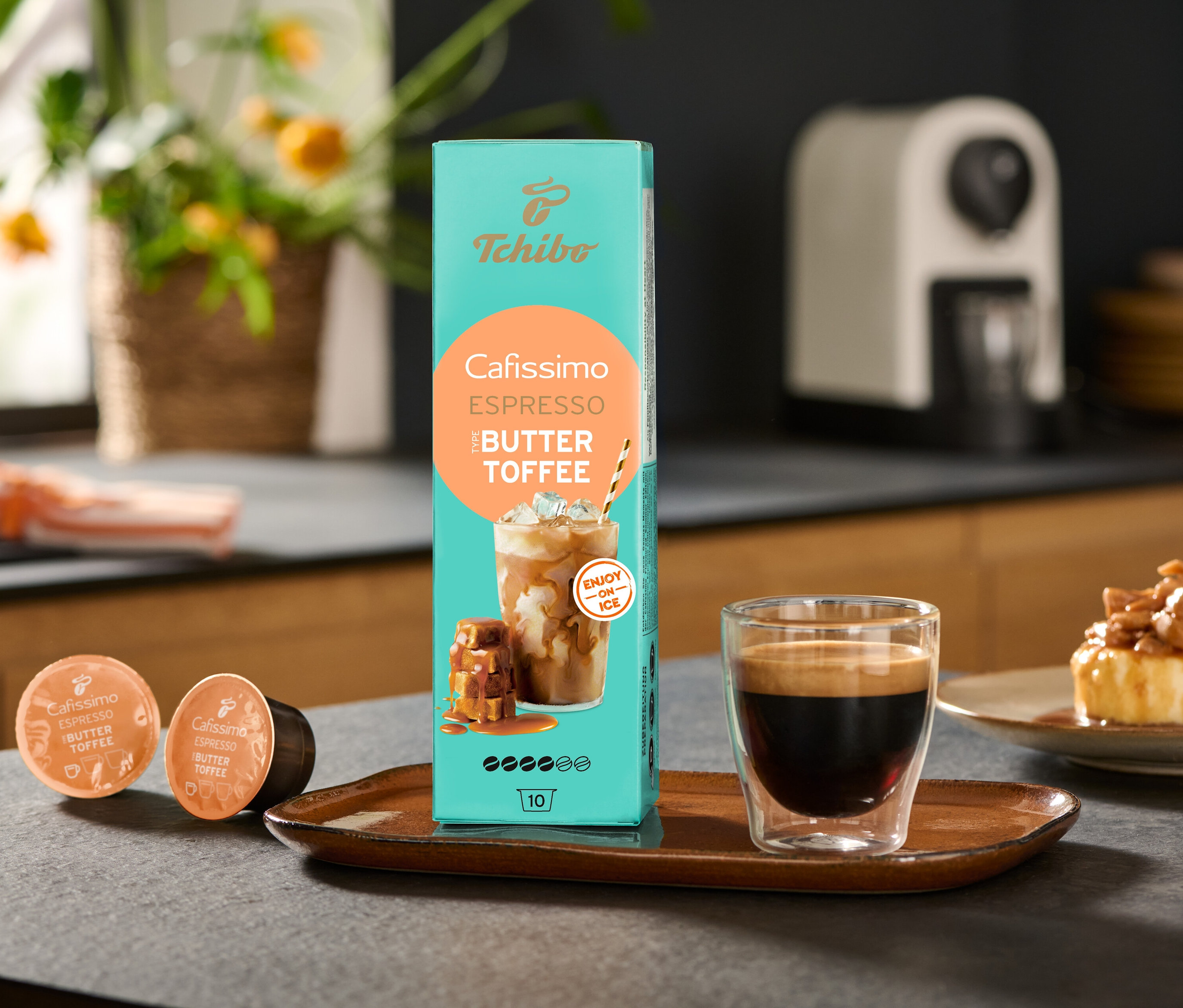 Egy csomag Cafissimo Flavoured Espresso – Buttertoffee és egy Cafissimo pure+, white kávéfőző áll az asztalon egy csésze kávé mellett.