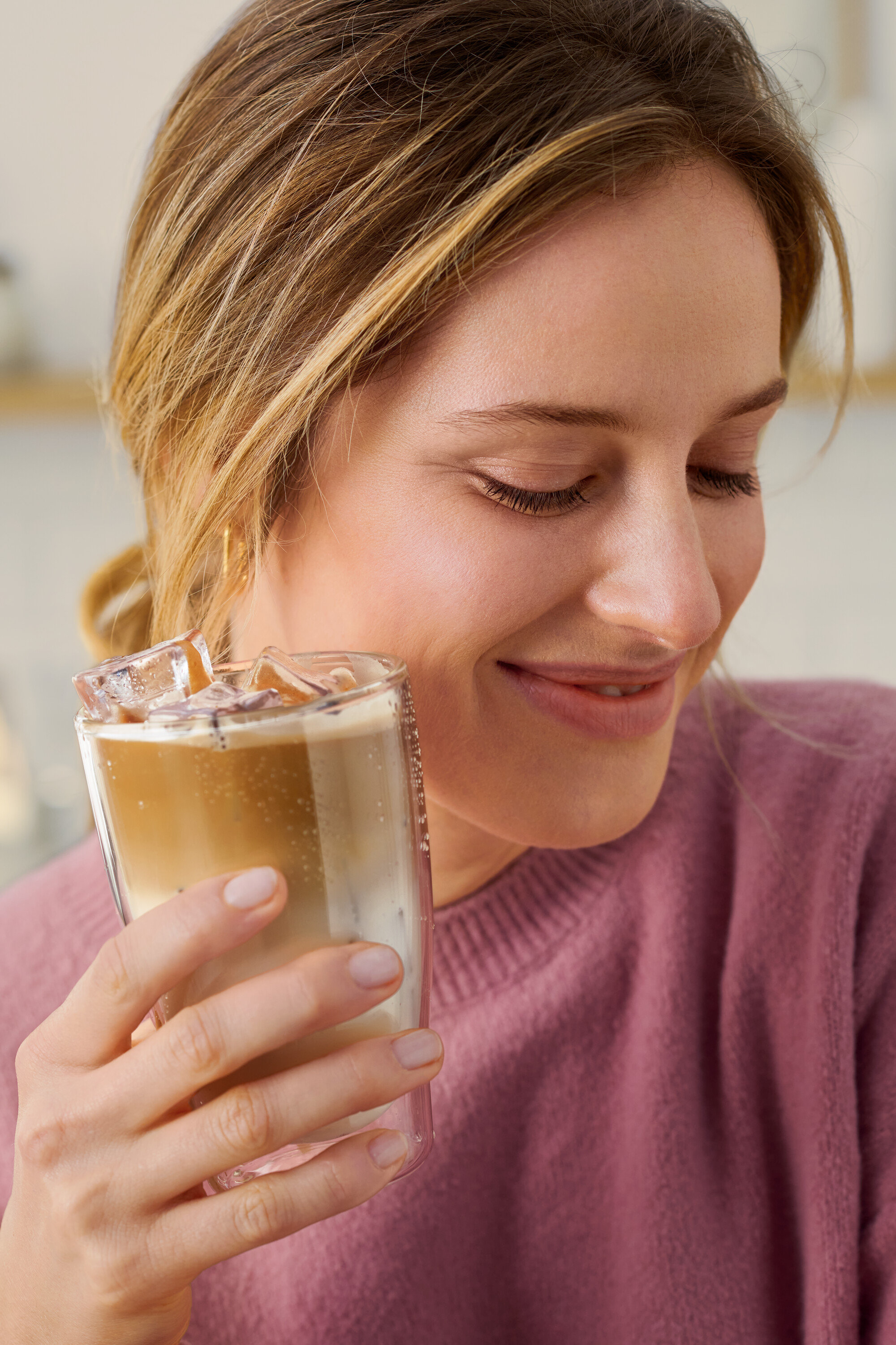 Nő tart egy poharat egy italral, ami jégkockákat tartalmaz. A háttérben egy fehér Cafissimo pure+ látható.
