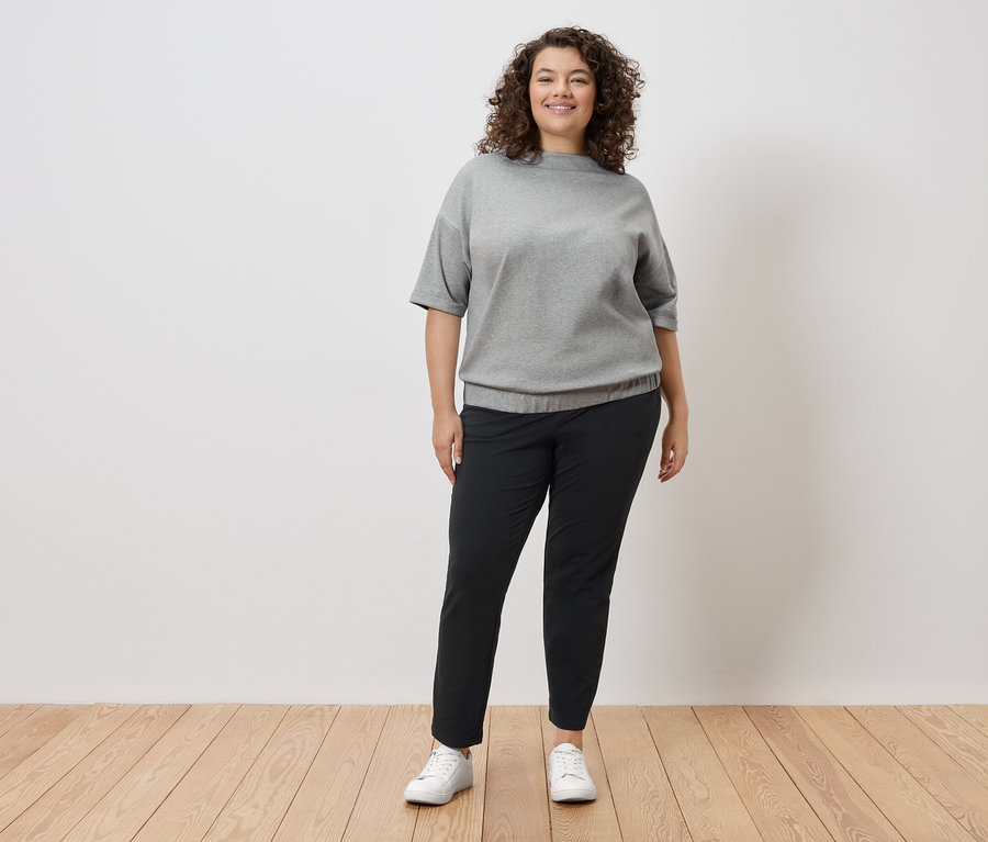 Egy göndör hajú nő áll egy fehér fal előtt, szürke pulóvert, fekete nadrágot és fehér tornacipőt visel.