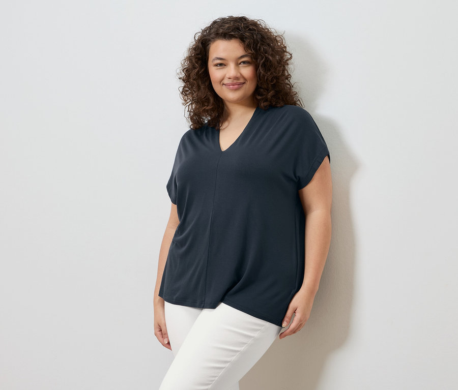 Egy göndör hajú nő áll egy fehér fal előtt, sötétkék pólót és fehér nadrágot visel.