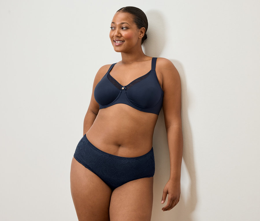 Nő sötétkék melltartóban és alsóneműben áll egy fehér falnál és mosolyog.