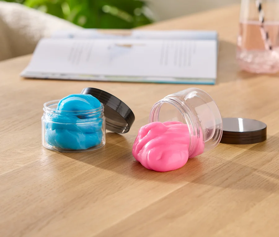 Két nyitott slime tartó edény egy fa asztalon; az egyik áll, benne kék slime, a másik az oldalán fekszik, rózsaszín slime ömlik ki belőle. A háttérben egy nyitott könyv és egy pohár rózsaszín folyadék látható.