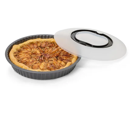 Egy kerek, bordázott, sötétszürke tortaforma sült almás pitével és mellette fekvő áttetsző fedéllel, fekete fogantyúval.