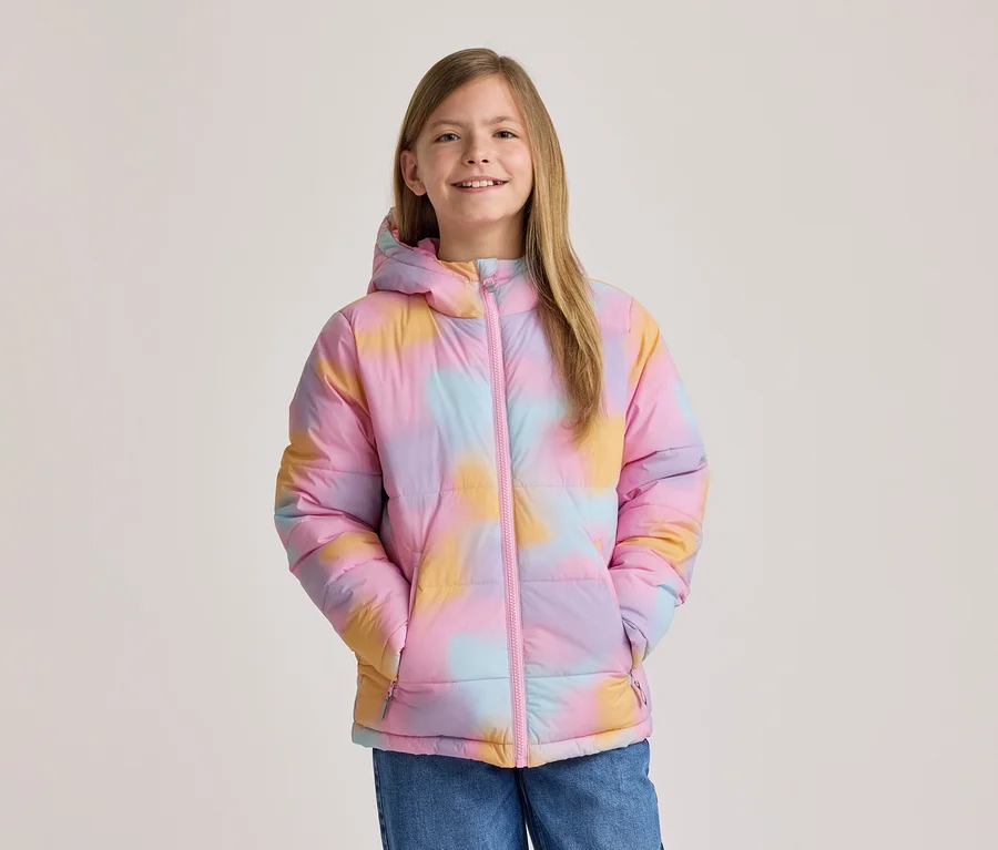 Mosolygó fiatal lány színes, rózsaszín, sárga és kék tie-dye mintás pufi kabátban, kapucnival és rózsaszín cipzárral, kezei a zsebében.