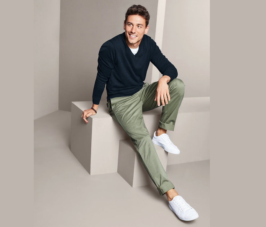 Egy férfi ül egy fehér kockán, sötétkék V-nyakú pulóvert, zöld nadrágot és fehér tornacipőt visel.