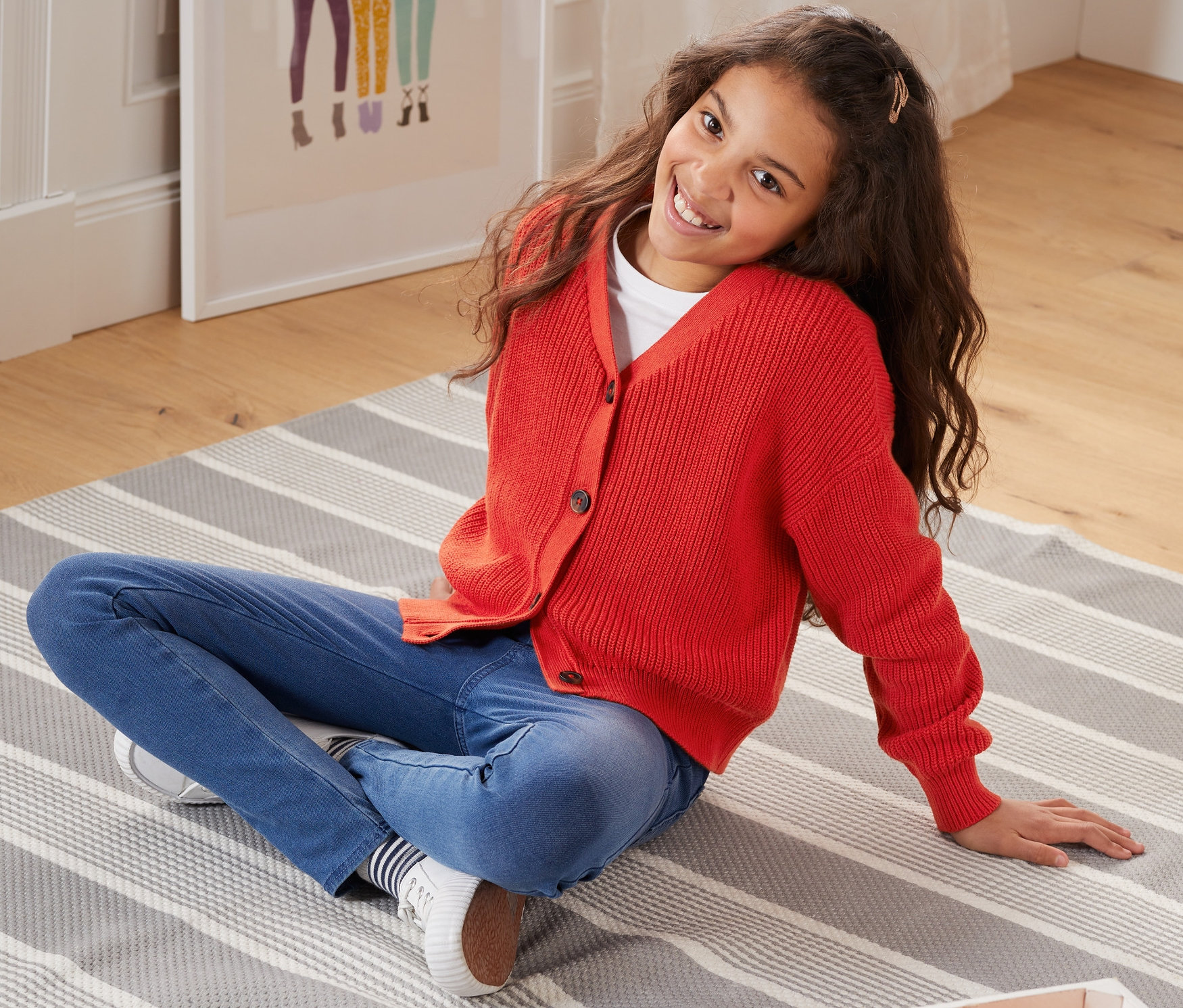 Egy lány ül egy csíkos szőnyegen, és egy piros kötött kardigánt visel.