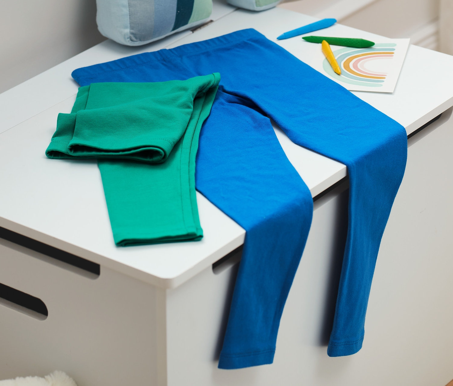 Egy fehér ládán kék és zöld leggingszek fekszenek, mellettük egy szivárvány dekoráció és színes ceruzák.