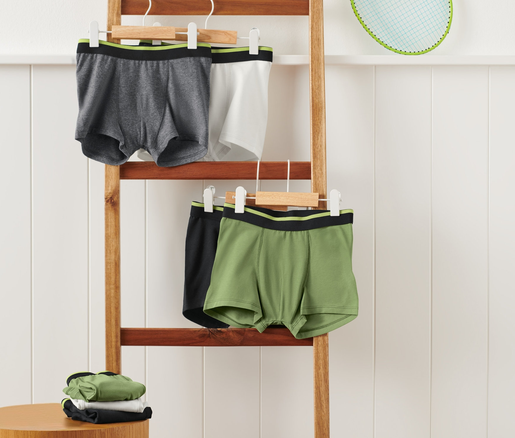 A falnak támasztott fa létrán szürke, fehér, fekete és zöld boxeralsók lógnak vállfán. Egy kis asztalon összehajtott zöld, fehér és fekete boxeralsók.