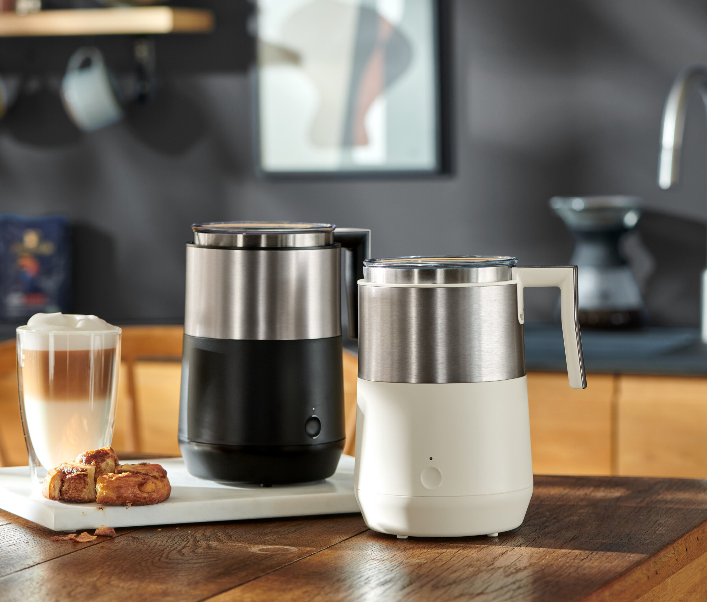 Két indukciós tejhabosító, egy fekete és egy krémfehér, egy fa asztalon áll egy lattés pohár és péksütemények mellett.