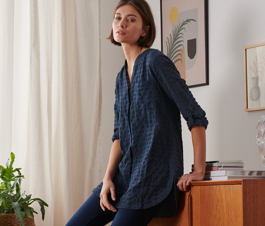 Egy nő egy fa komódon ül, sötétkék szövött tunikát és kék leggingset visel.