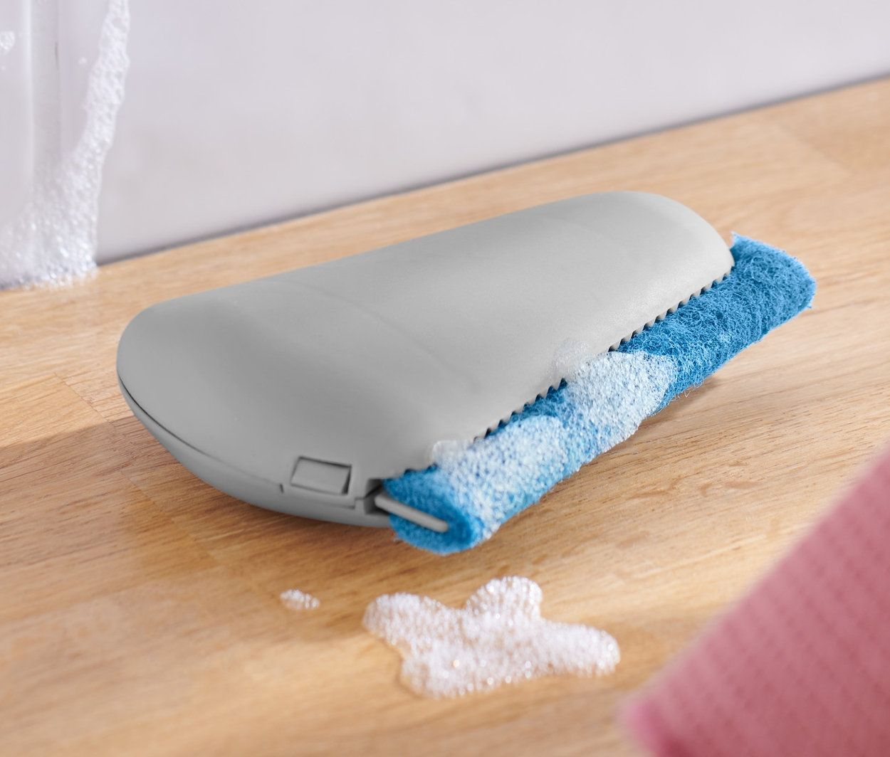 Egy él- és fugatisztító egy fa felületen fekszik egy fehér tál és egy rózsaszín kendő mellett.