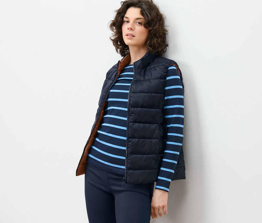 Nő a falnak támaszkodva, sötétkék/barna női kifordítható steppelt mellényt, csíkos pólót és nadrágot visel.